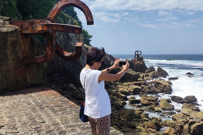 Person taking a photo of a rusty metal sculpture by the ocean on a rocky coastline.