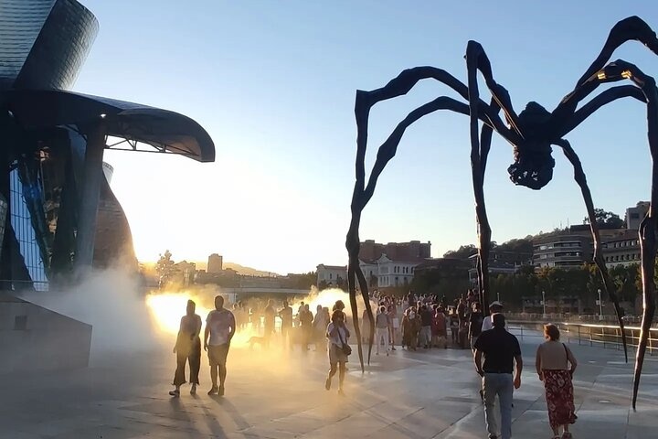 People walking near a large spider sculpture with mist and cityscape at sunset.