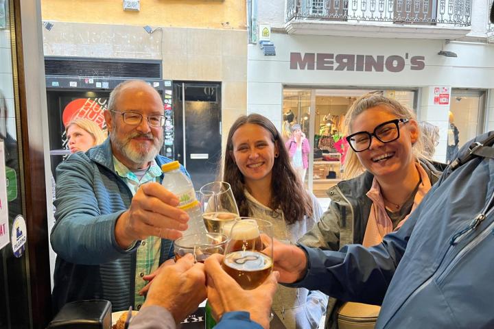 Three people smiling and toasting with drinks in a bar with a street view window.