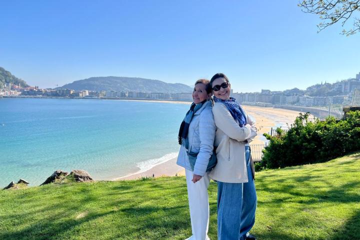 Two people smiling on a grassy hill by the ocean on a sunny day.