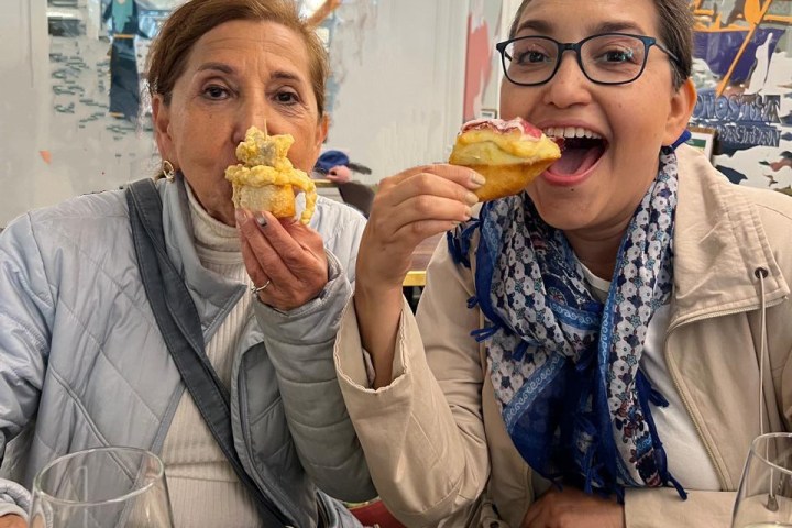 Two women smiling while eating pastries indoors, with wine glasses on the table.