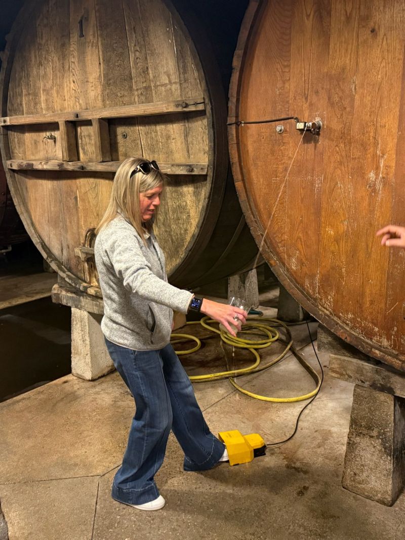 Two people inspecting large wooden barrels in a cellar, one holding a glass.
