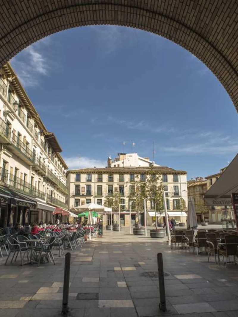 View of a plaza with cafes and old buildings under a brick archway on a sunny day.