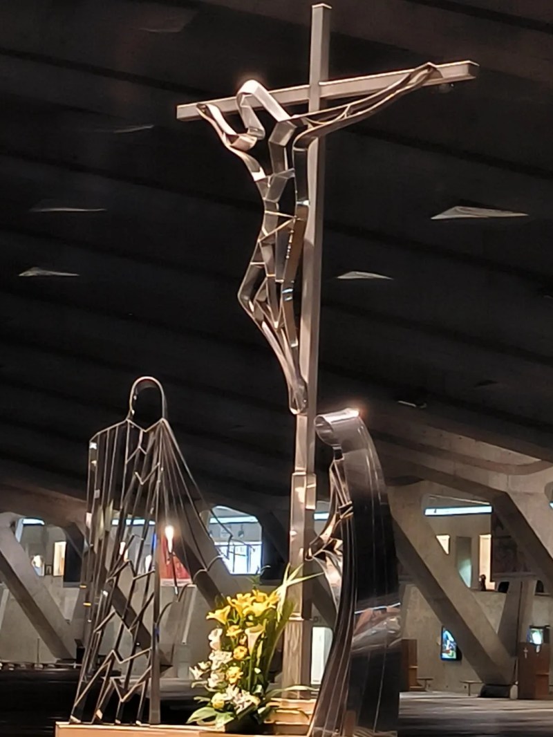 Modern metal crucifix sculpture with abstract figures and flowers in a church setting.
