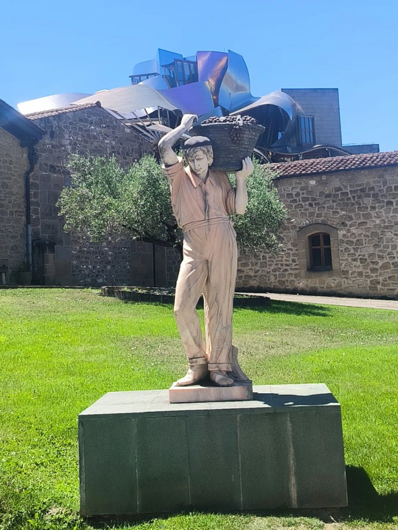 Stone statue of a man carrying a basket on his shoulder, in front of a building with modern architecture.
