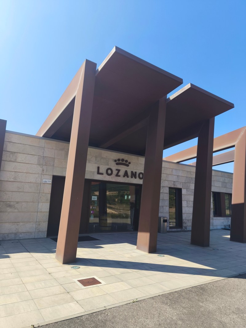 Entrance of a modern building with tall brown beams and 'LOZANO' sign on a sunny day.