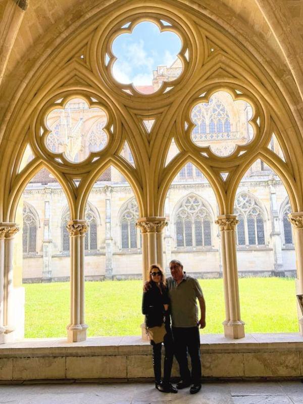 Two people stand in a Gothic cloister with ornate arches overlooking a grassy courtyard.