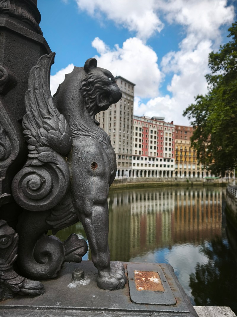 Winged lion sculpture on a bridge with colorful buildings and canal in the background.