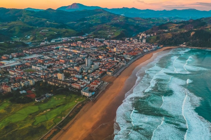Aerial view of a coastal city with beach and waves, surrounded by green hills.