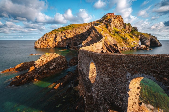 Stone bridge leads to rocky island with a small red-roofed building, surrounded by sea and clouds.