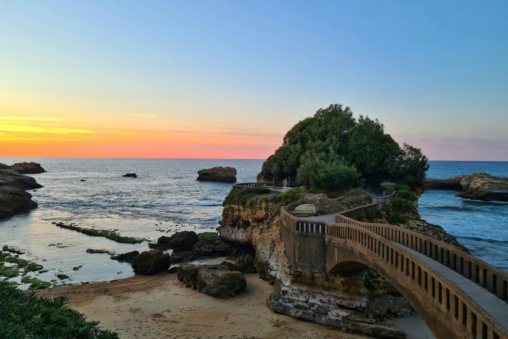 Sunset over ocean with small bridge leading to rocky island, sandy beach, and greenery.