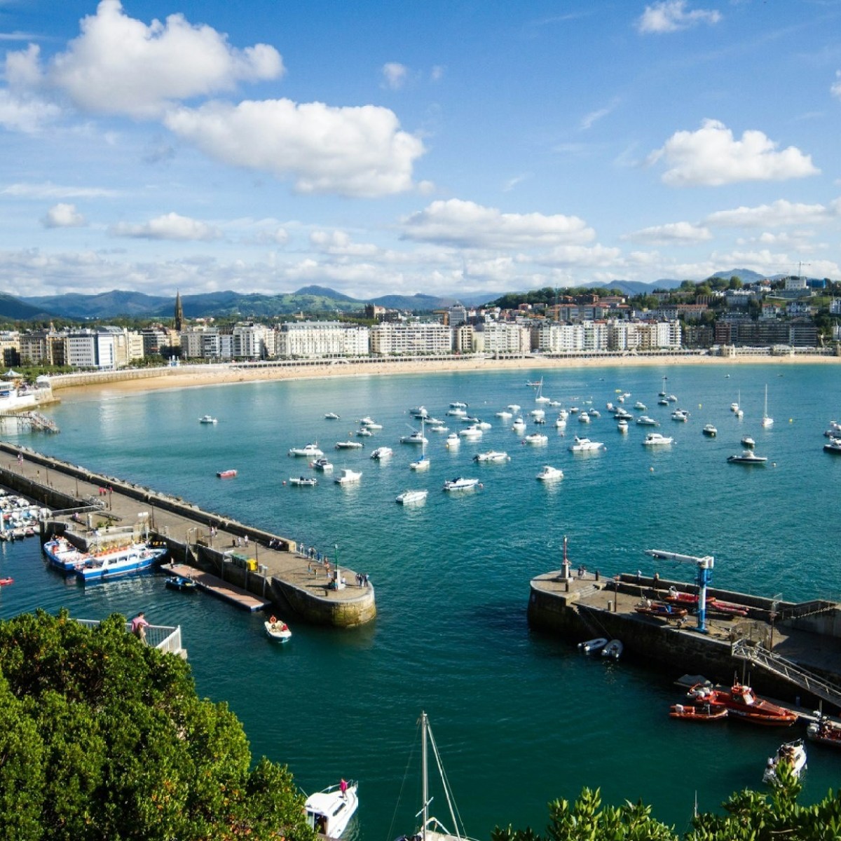 A scenic bay with boats, a pier, and city buildings under a partly cloudy sky.