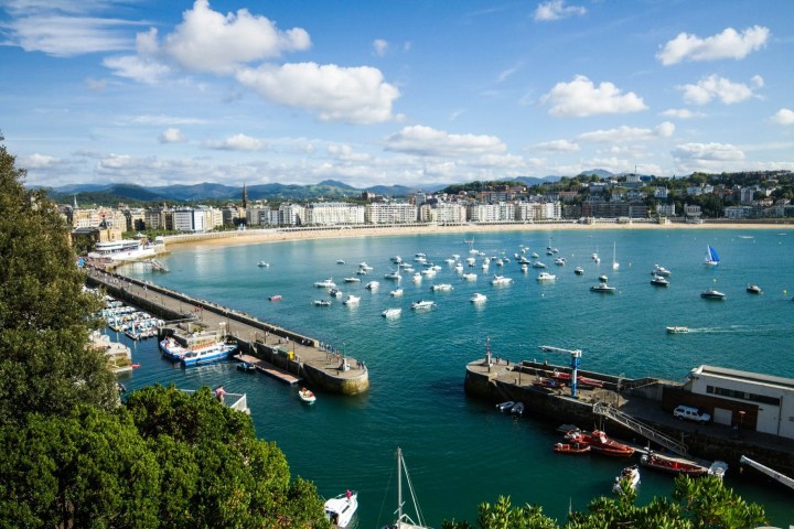 A scenic bay with boats, a pier, and city buildings under a partly cloudy sky.