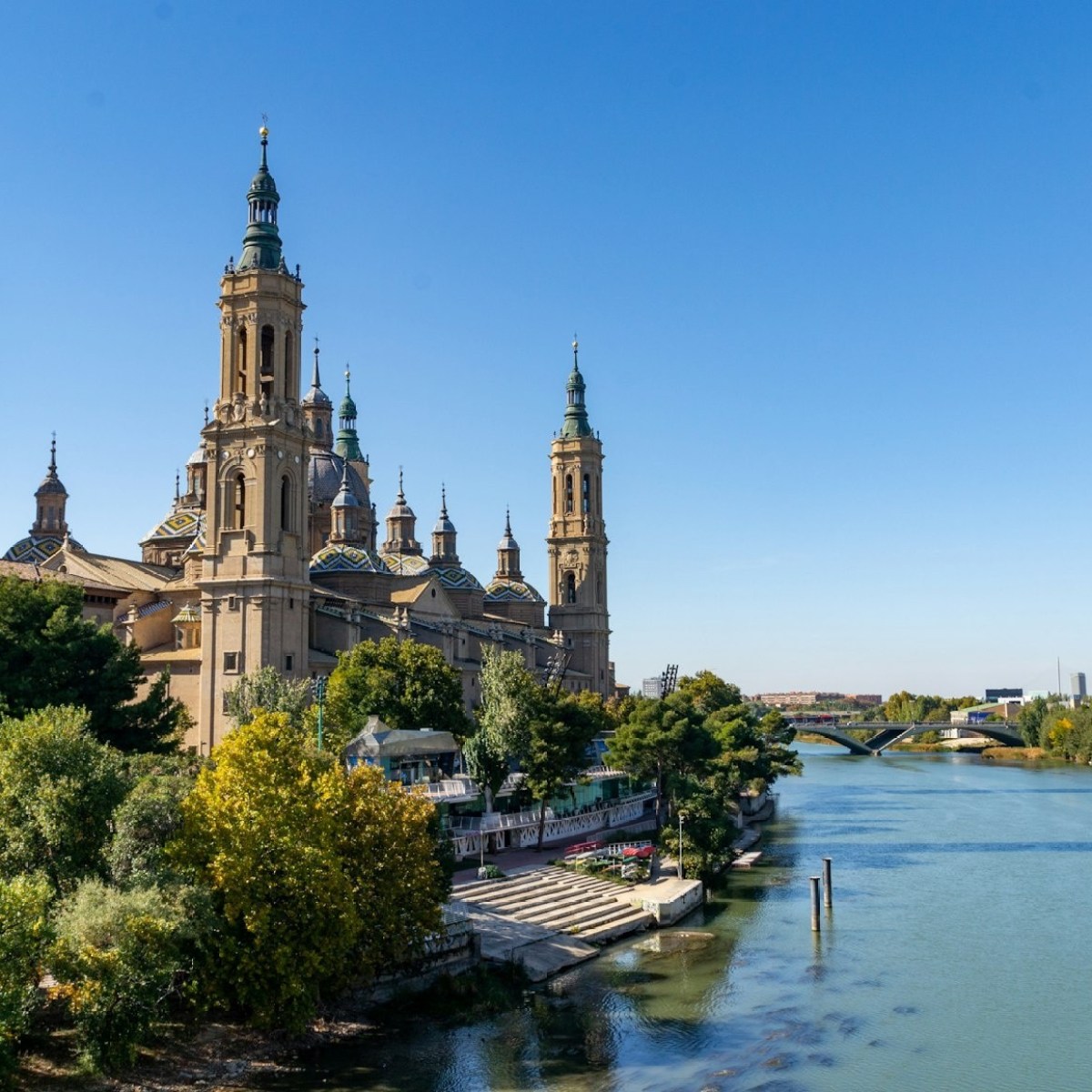 Basilica by a river, surrounded by trees under clear blue sky.