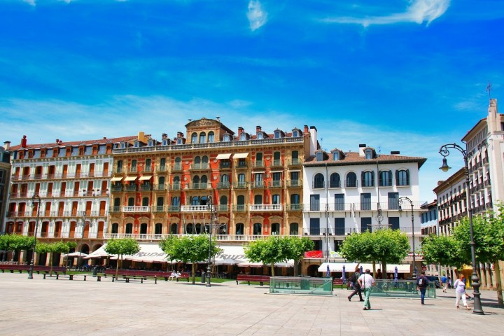 Colorful buildings under blue sky in a city square with trees and people walking.