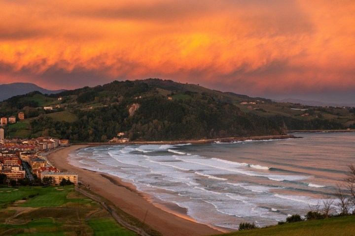 Beach with waves near town and hills under vibrant orange sky at sunset.