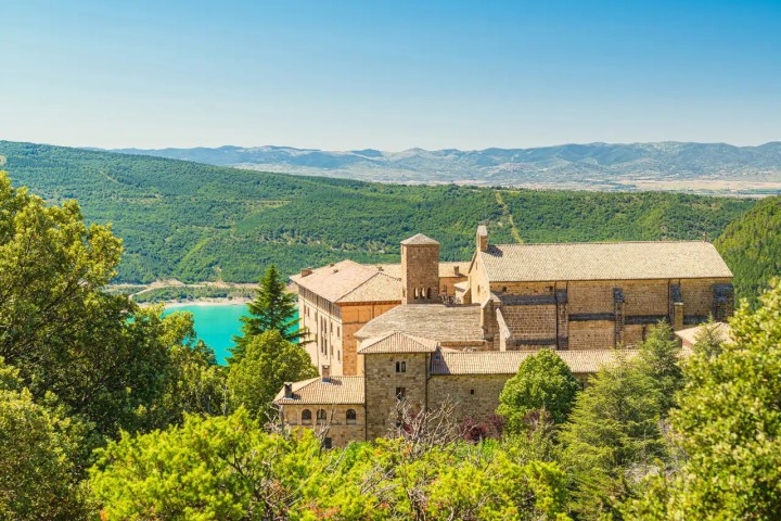 Historic stone building surrounded by lush trees with hills and a lake in the background.